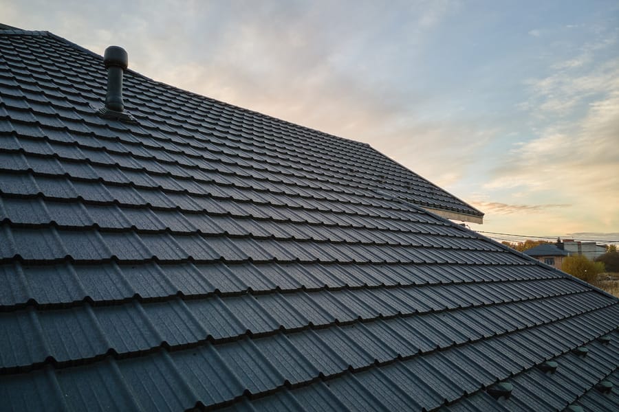 Close-up of a stone coated steel roofing system featuring charcoal grey textured panels and a plumbing vent stack against a sunset sky.